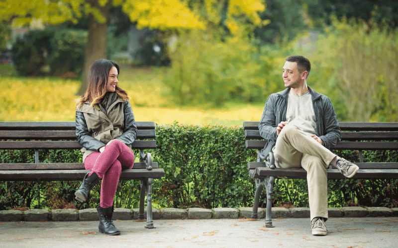 casal sentados em banco de jardim separados se olhando e sorrindo um para o outro.